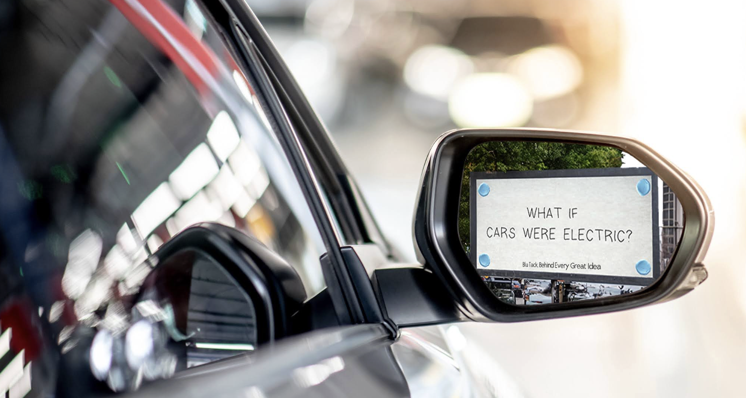 Car side mirror with an electronic sign inside that reads, 'What if cars were electric?' with a background of trees and a bright sky.