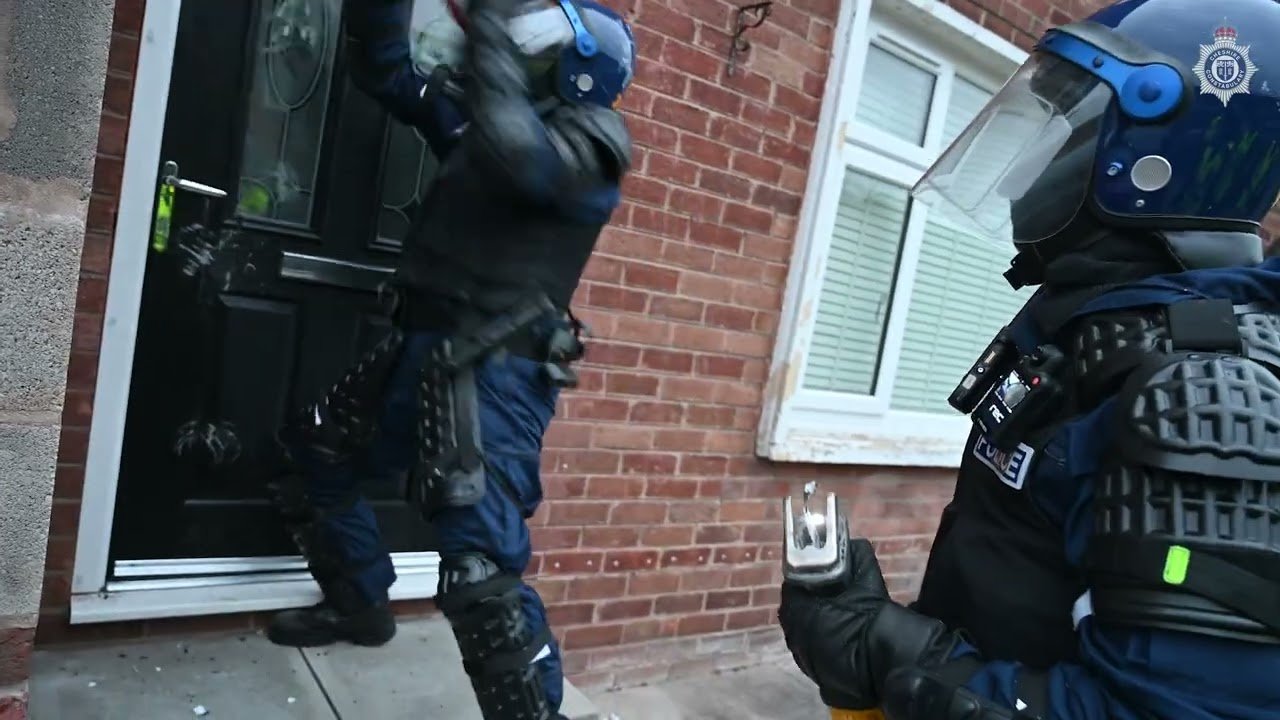 Two police officers in riot gear outside a brick house, with one opening the door and the other holding a mobile phone, wearing helmets and protective vests.