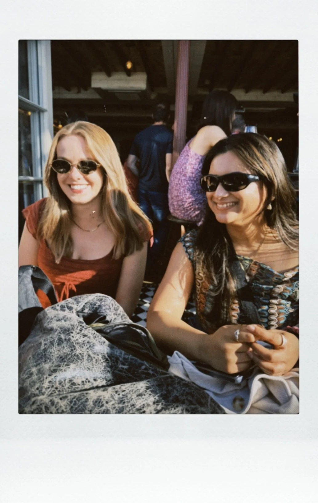 Two women smiling and wearing sunglasses, sitting at a table in a restaurant or cafe, with people in the background.