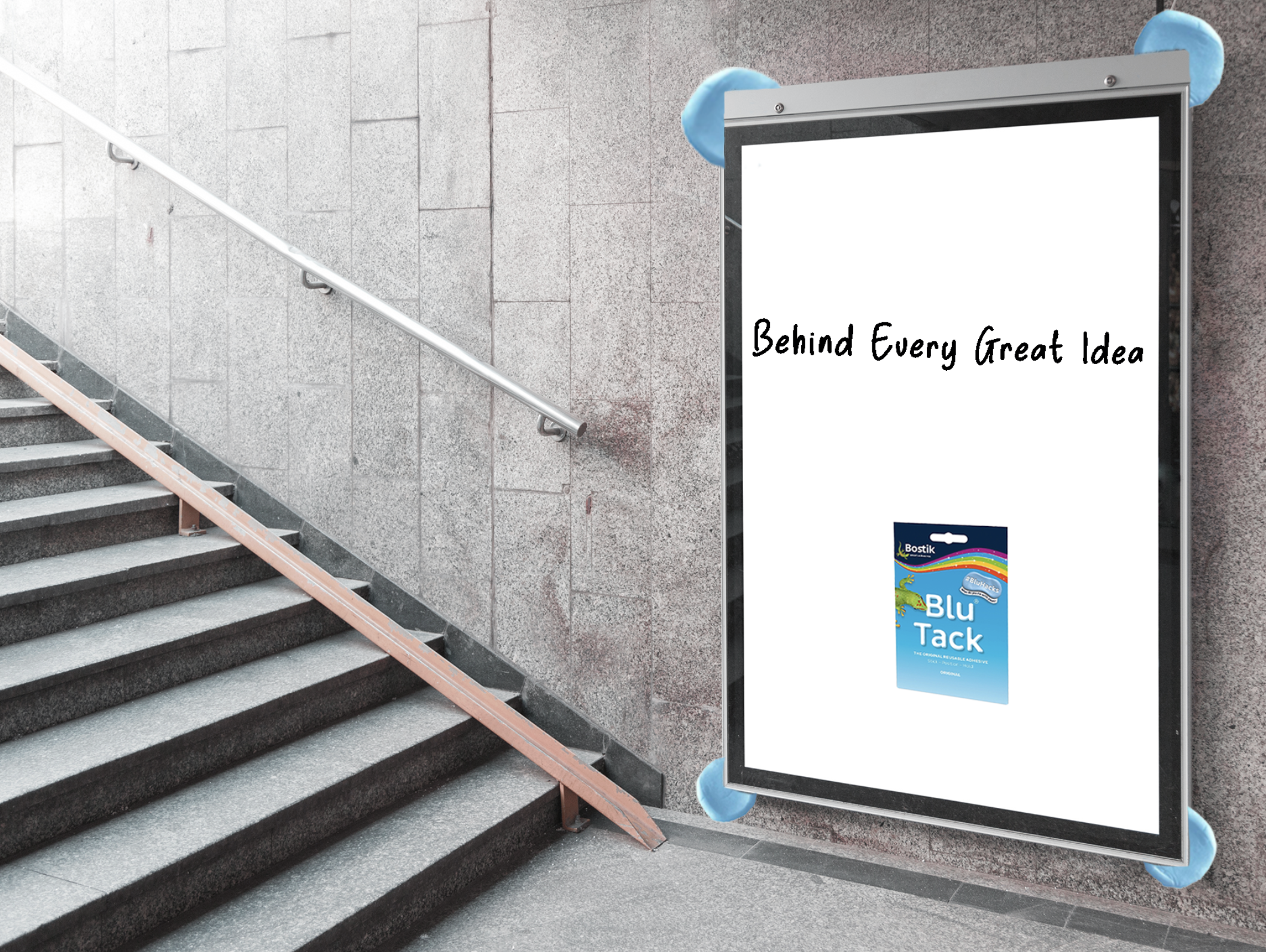 Outdoor staircase leading up with a metal handrail on the left side. To the right, there is a large, empty billboard frame with blue protective padding on the corners. The ground and wall are made of gray stone tiles. The billboard displays the phrase "Behind Every Great Idea" in black cursive font and a blue advertisement for Blu Tack adhesive at the bottom.