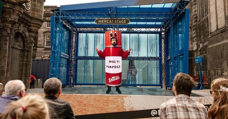 A person dressed in a Mr. Sheen multi-purpose cleaning spray can costume performing on an outdoor stage labeled 'Mercat Stage' with an audience watching.