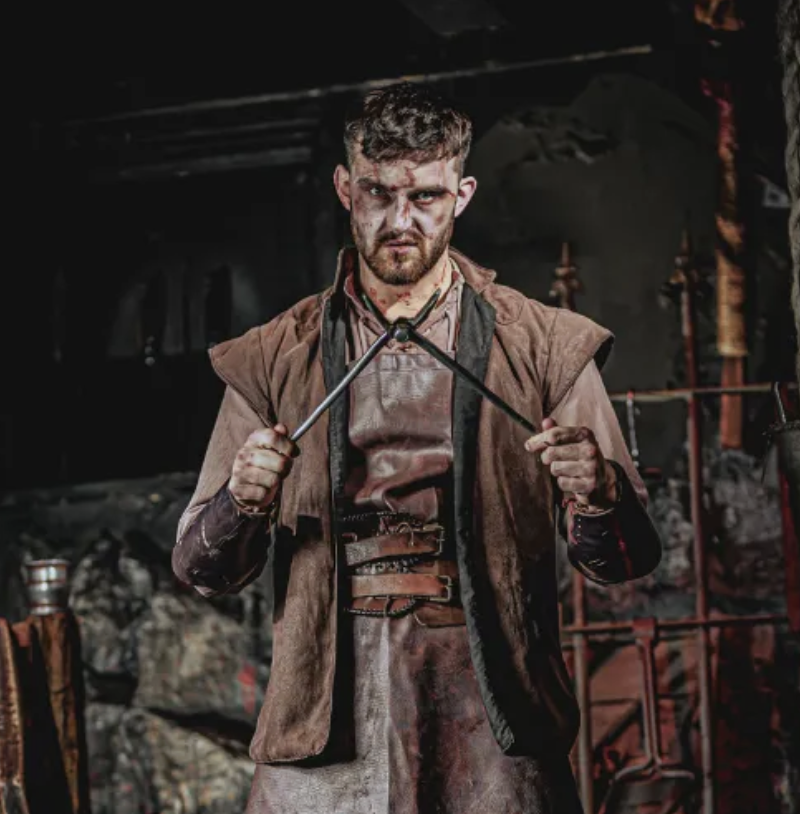 A man with a rugged appearance and messy hair holding two metal rods, standing in a dark, fiery blacksmith workshop with tools and rocks in the background.