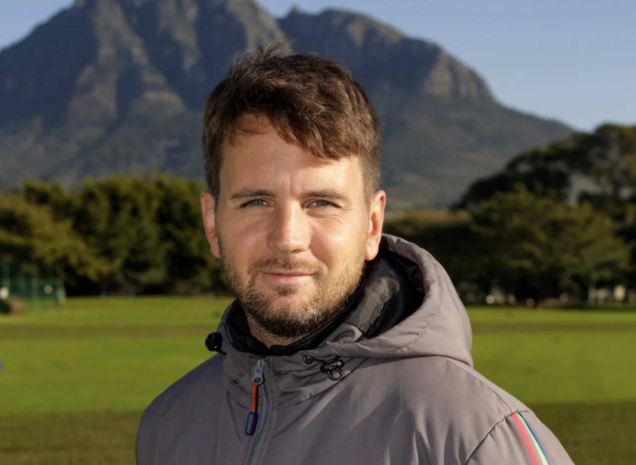 A man outdoors with a mountain and trees in the background, wearing a gray jacket with a colorful stripe on the sleeve.
