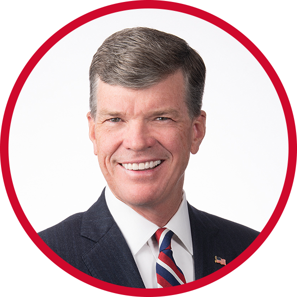 Portrait of a smiling man wearing a dark suit, white shirt, and striped tie, with an American flag lapel pin, against a white background with a red circular border.