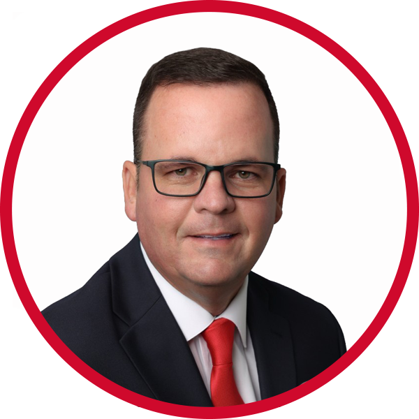 A professional man wearing a dark suit, white shirt, and red tie, with black glasses, posing against a white background with a red circular border.