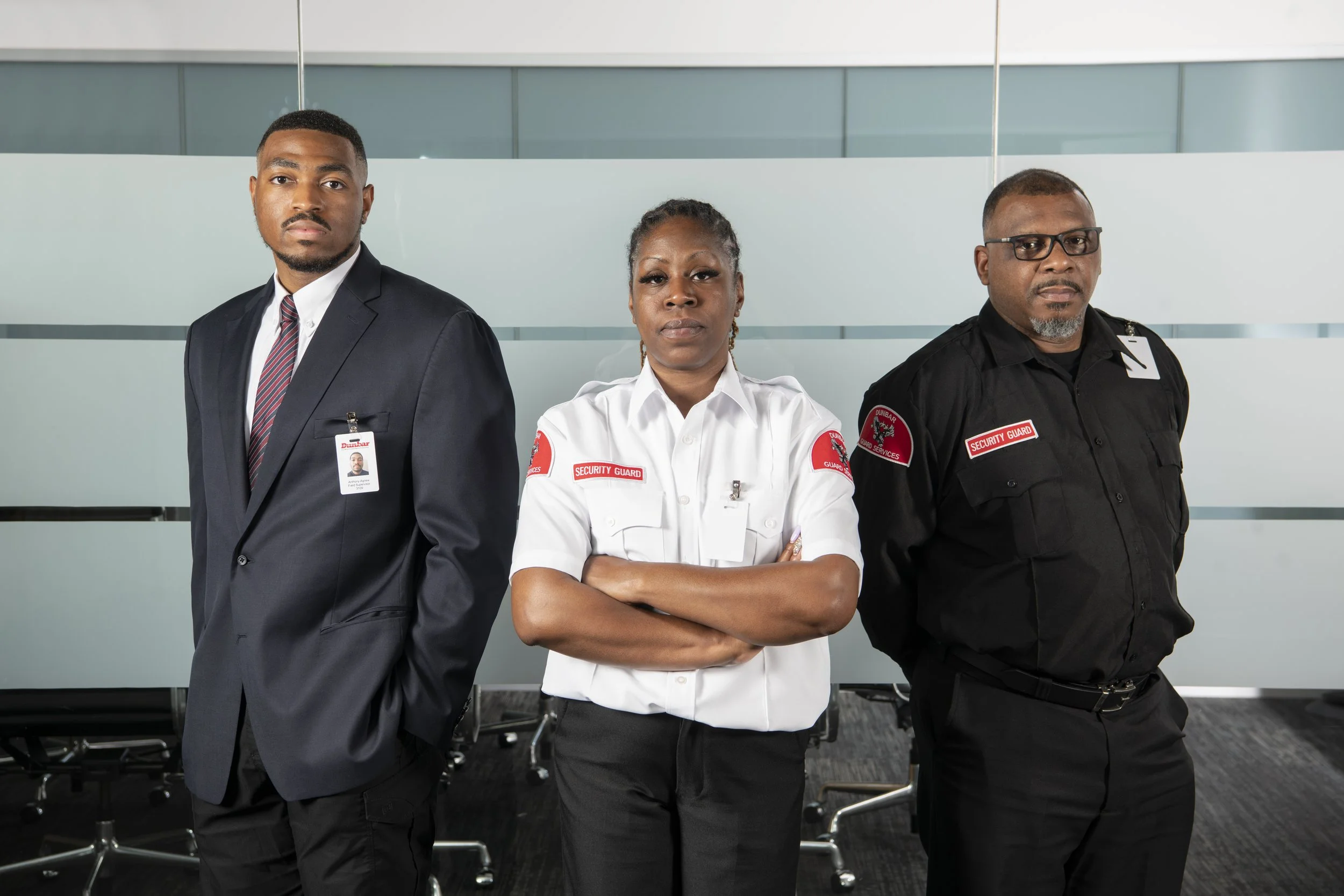 Three security personnel standing in a row in an office environment. The man on the left is wearing a dark suit and tie with an ID badge, the woman in the middle is wearing a white security uniform with her arms crossed, and the man on the right is wearing a black security uniform with glasses. They all face forward with serious expressions.