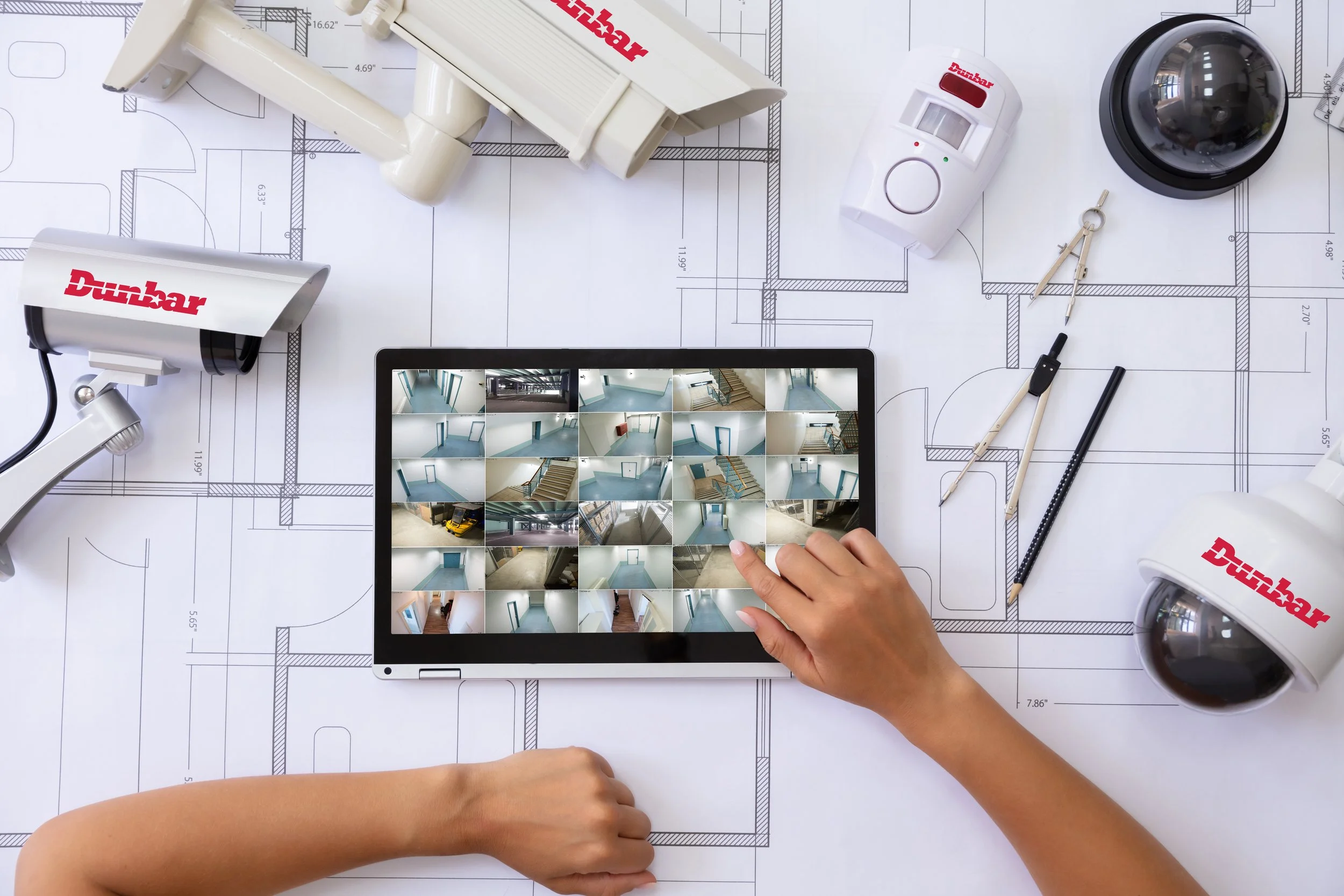 Person using a tablet to view images of a room interior on a blueprint spread out on a table with various tools and security cameras.