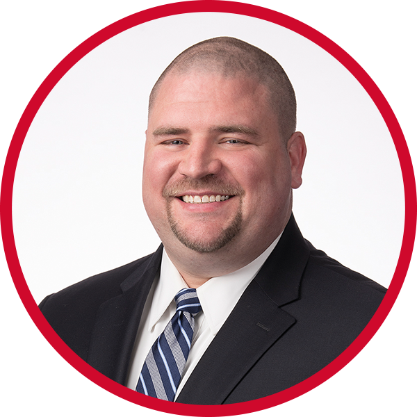 A professional headshot of a man with a shaved head, goatee, wearing a black suit, white shirt, and striped tie, smiling in front of a white background with a red circular border.