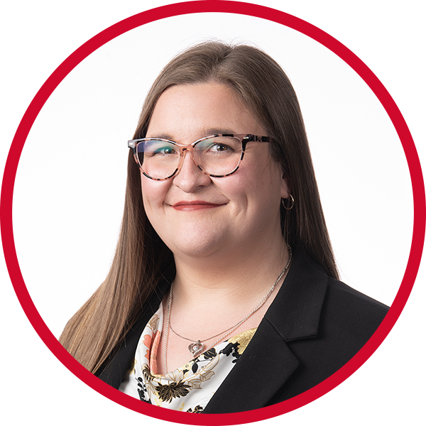 Professional woman with long brown hair, glasses, wearing a black blazer and floral blouse, smiling against a red and white circular background.