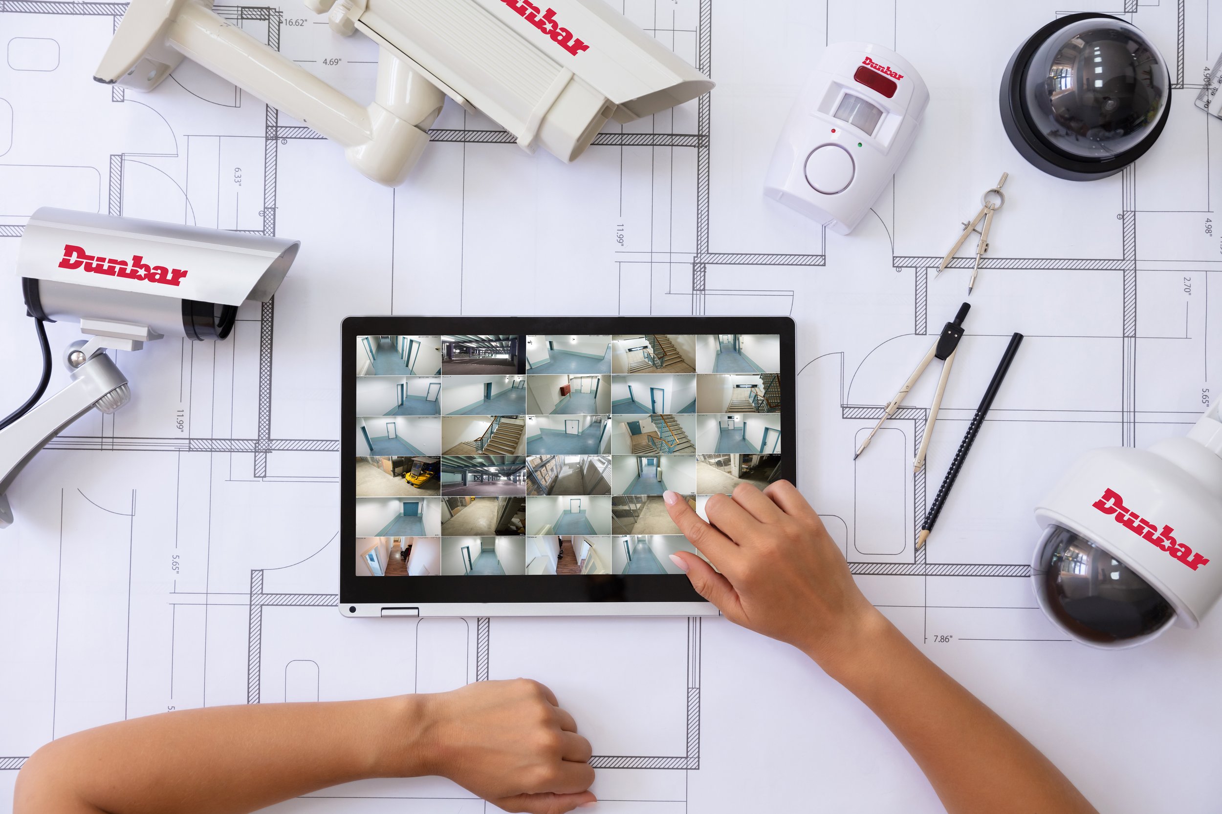 Top-down view of a person's hand holding a tablet showing multiple room images, surrounded by construction tools, security cameras, a motion detector, a compass, and architectural blueprints on a table.