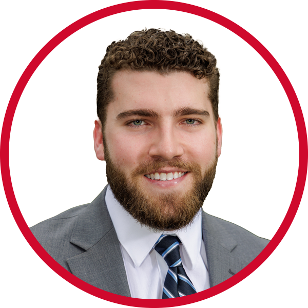 Portrait of a young man with curly brown hair and a beard, smiling, wearing a gray suit and a striped tie, against a red background.