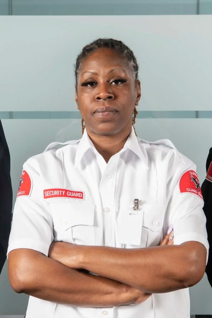 A woman in a security guard uniform with crossed arms standing in front of a light-colored wall.