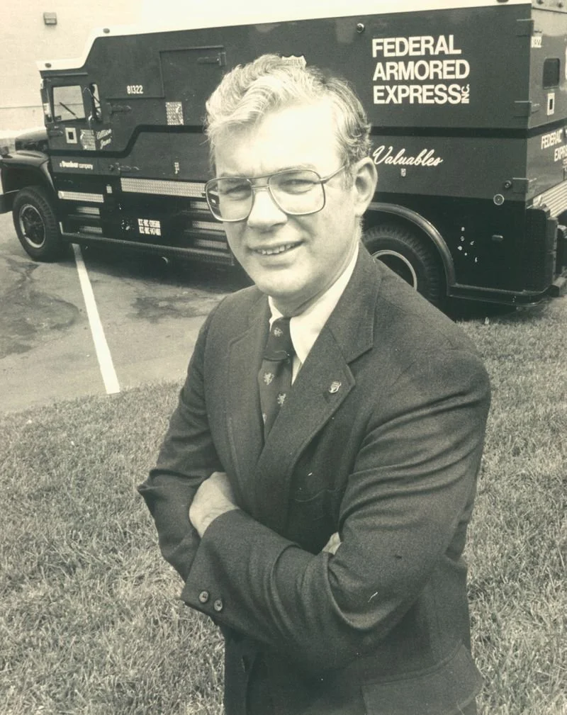 A man wearing glasses, a suit, and a tie with a military pin, standing outdoors with arms crossed, in front of a vehicle marked 'Federal Armored Express' and a building.