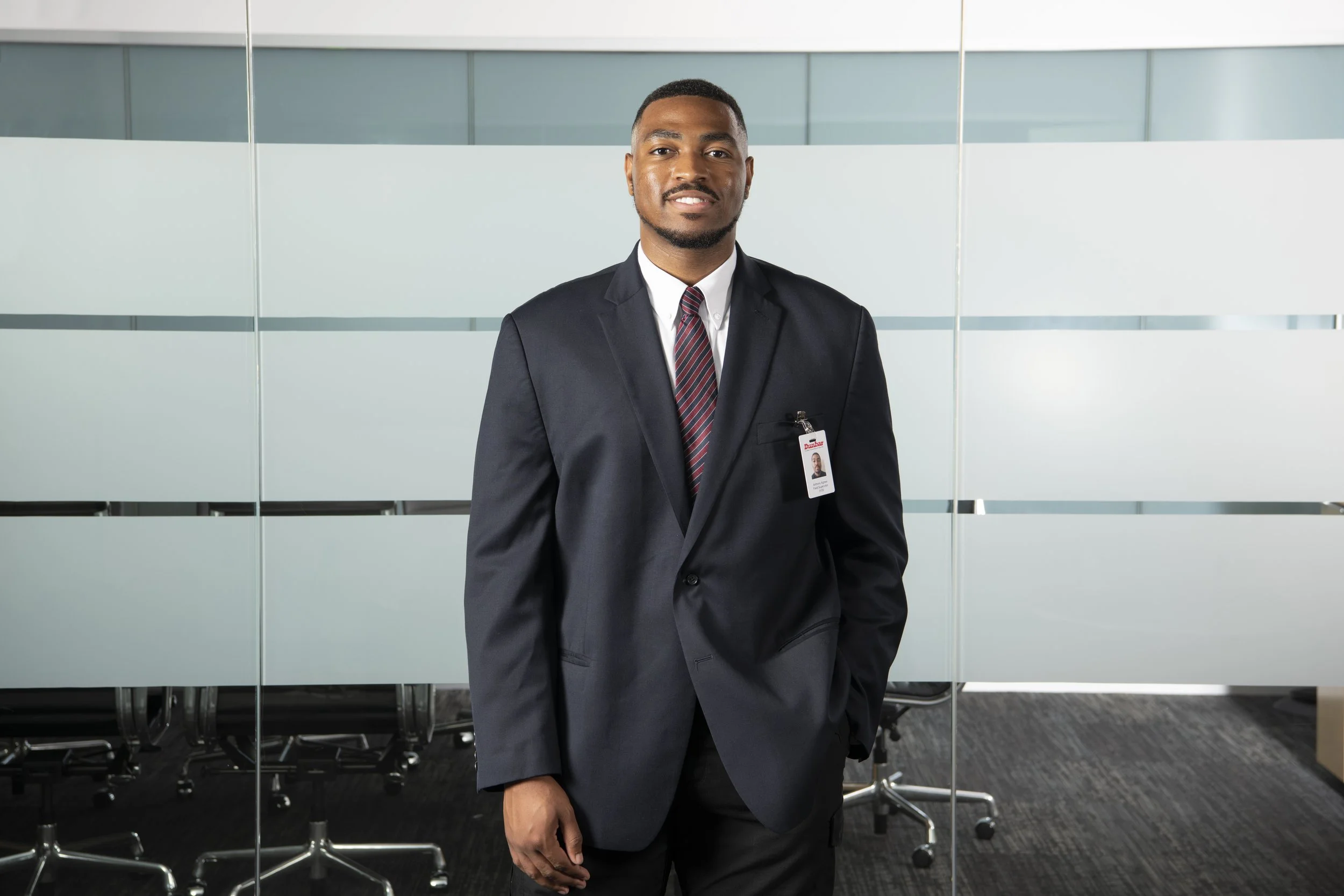 A man in a dark suit with a white shirt and striped tie standing in an office conference room with glass walls and empty chairs.