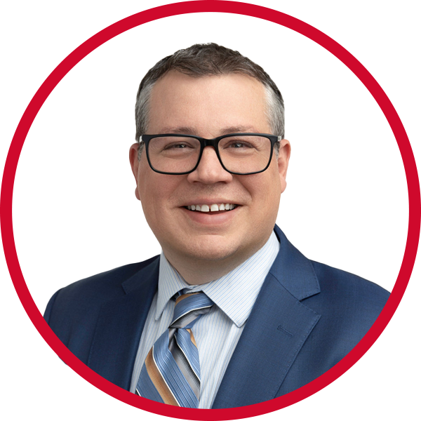 Portrait of a smiling man wearing glasses, a dark blue suit, a light blue shirt, and a striped tie, against a red and white background.