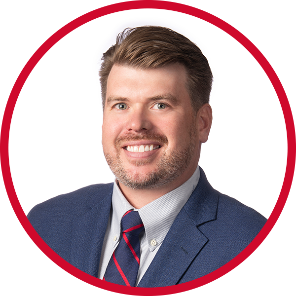 Professional headshot of a man with brown hair and a beard, wearing a navy blue suit, light gray shirt, and a striped red and navy blue tie, against a white background with a red circular border.