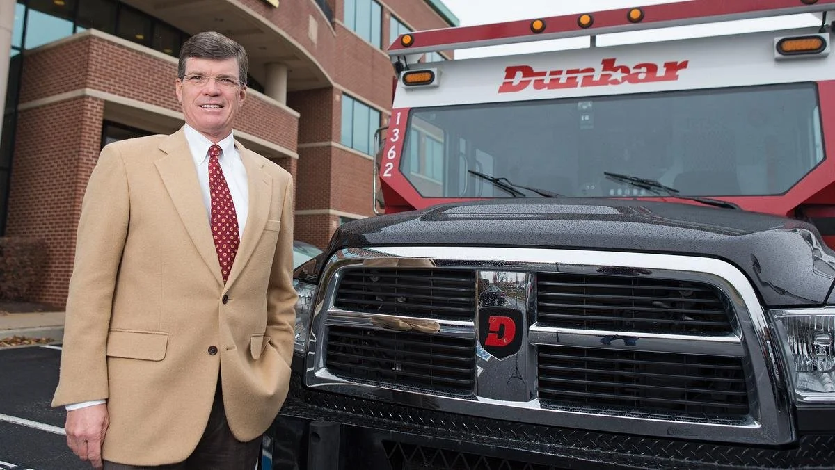 A man in a beige suit with a red tie standing next to a black truck with a Dodge logo on the front grille, in front of a brick building and a Dunkin' truck.