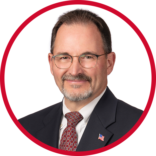 A man with glasses, a beard, and mustache, wearing a dark suit, white shirt, a red patterned tie, and a pin of the American flag on his lapel, smiling against a white background with a red circular border.
