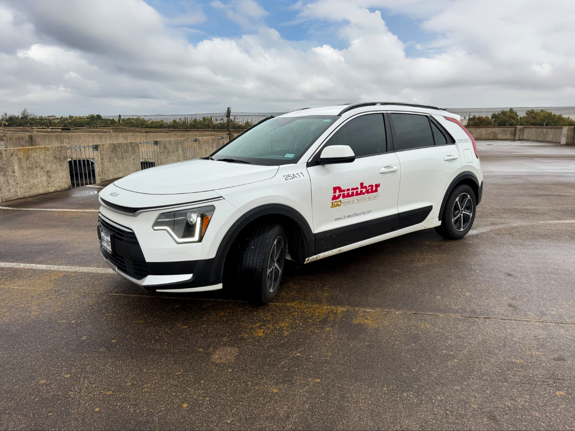 A white security vehicle parked in an empty parking lot under cloudy skies, with a concrete barrier and a landscape in the background.