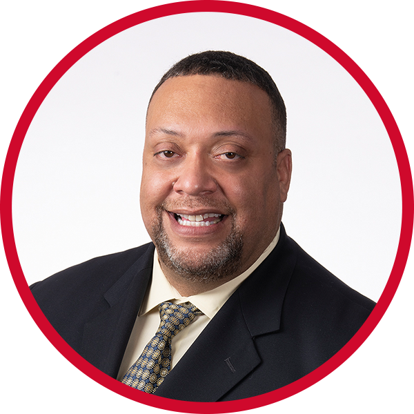 Professional headshot of a smiling man wearing a dark suit, light-colored shirt, and patterned tie, against a white background with a red circular border.