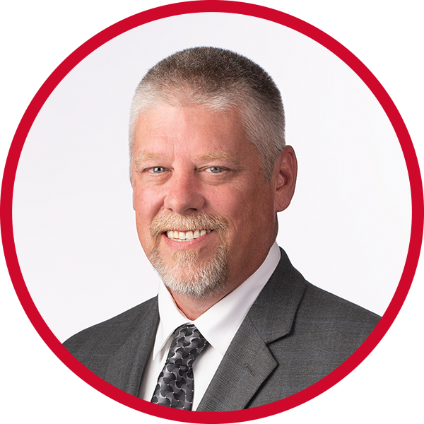 Headshot of a middle-aged man with short gray hair, a beard, wearing a gray suit, white shirt, and patterned tie, smiling against a white and red background.