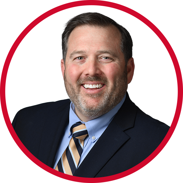 A smiling man in a dark suit, blue dress shirt, and striped tie, with short brown hair and a beard, against a white background with a red circular border.