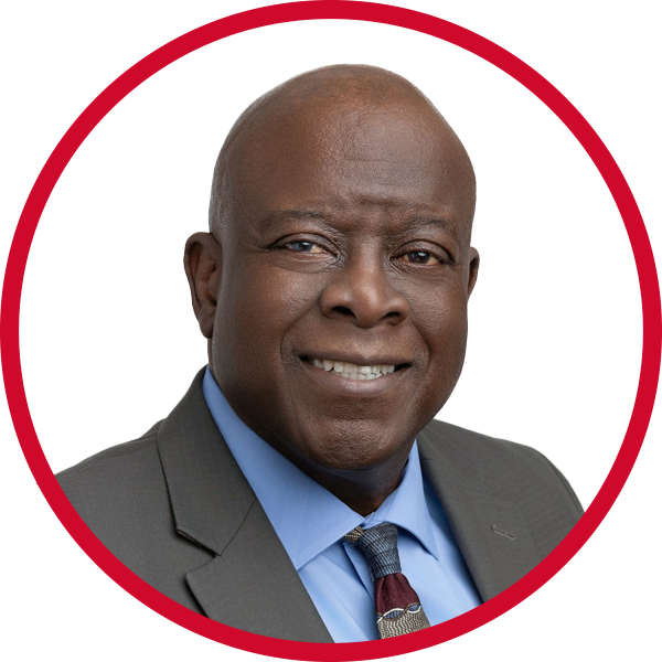Professional headshot of a middle-aged Black man wearing a grey suit, blue shirt, and patterned tie, smiling against a white and red background.