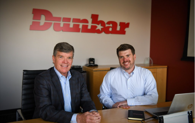 Two men sitting at a conference table in an office with a large Duralar logo on the wall behind them. They are smiling at the camera, and there is a laptop and a notebook on the table.