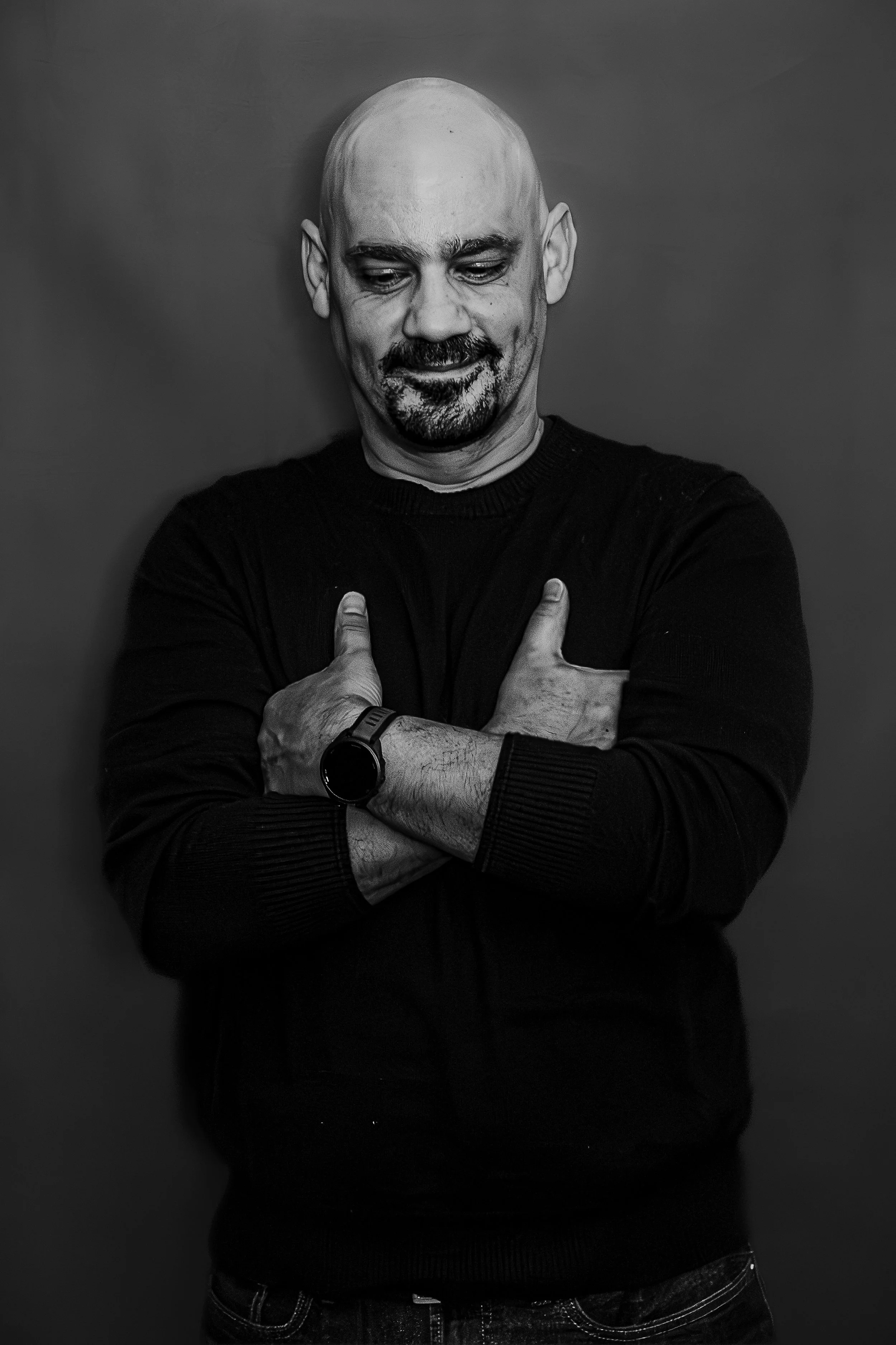 A black-and-white self-portrait by Joseph Sharp, photographer of architecture, interiors, portraits, nature, and street scenes, capturing a quiet moment of personal reflection with folded arms, a subtle smile, and a minimalist backdrop.