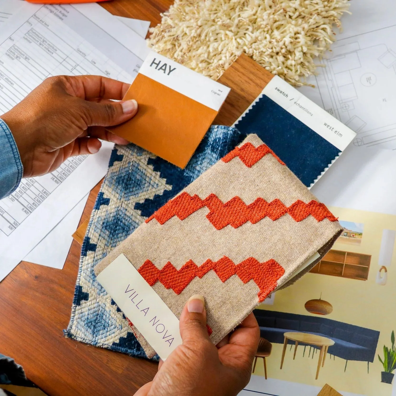 Person holding fabric swatch samples over a wooden table with fabric and architectural plans. Three fabric samples include brown, navy, and beige with orange zigzag embroidery. The person's hand is visible, and they appear to be examining the samples for interior design or renovation purposes.