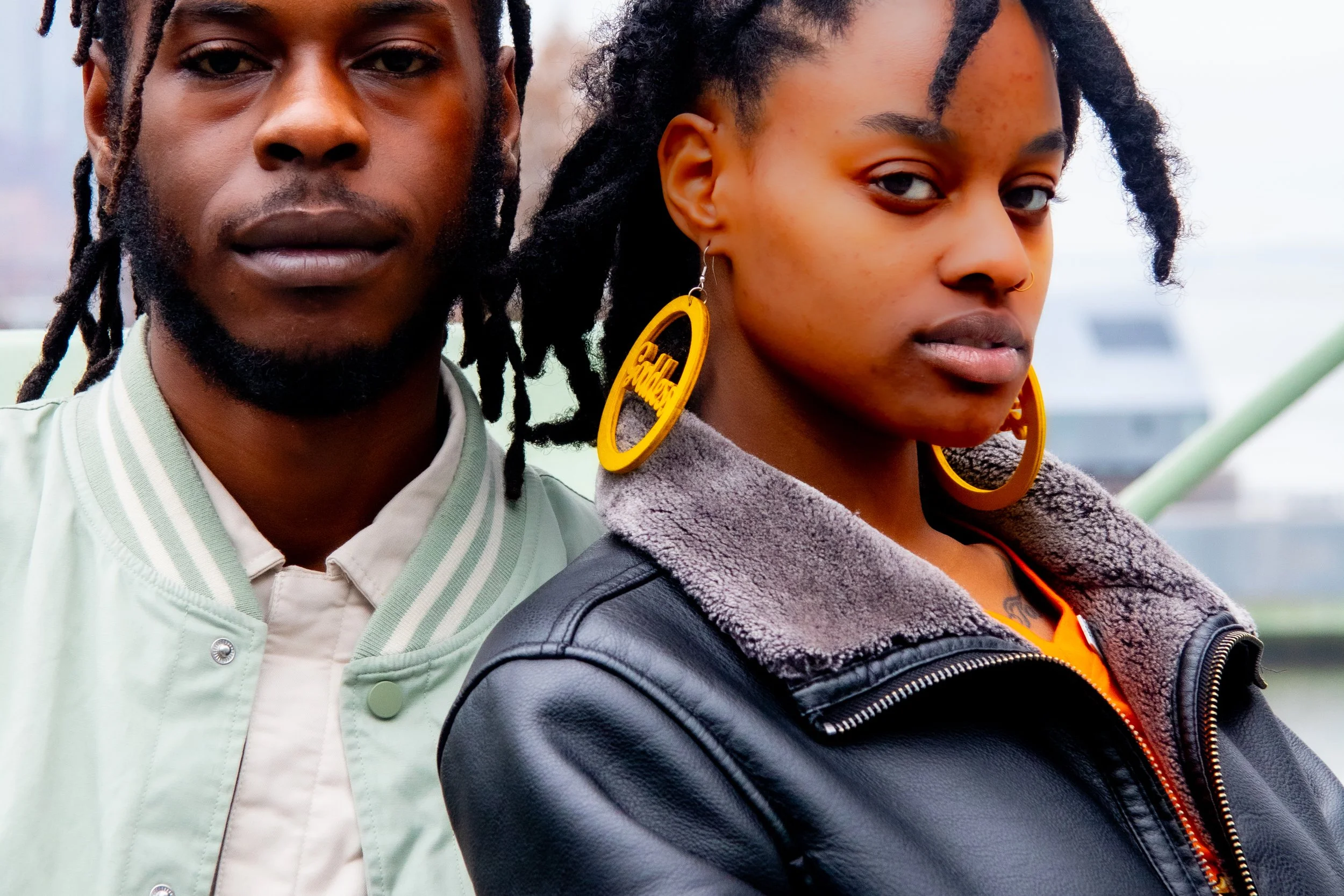 Close-up of a young Black man and woman standing outdoors, with the man slightly behind the woman. Both are looking at the camera, with the woman wearing large yellow earrings and the man wearing a light-colored jacket.