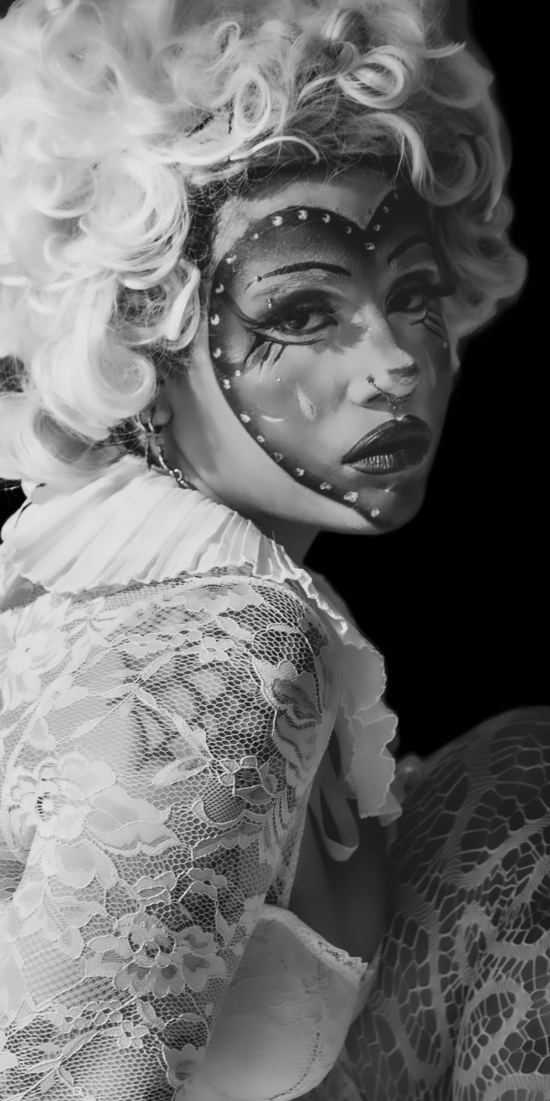 A person with elaborate face makeup, curly blonde hair, and wearing a lace outfit. The makeup includes artistic designs with rhinestones and tears, and the photo is in black and white.