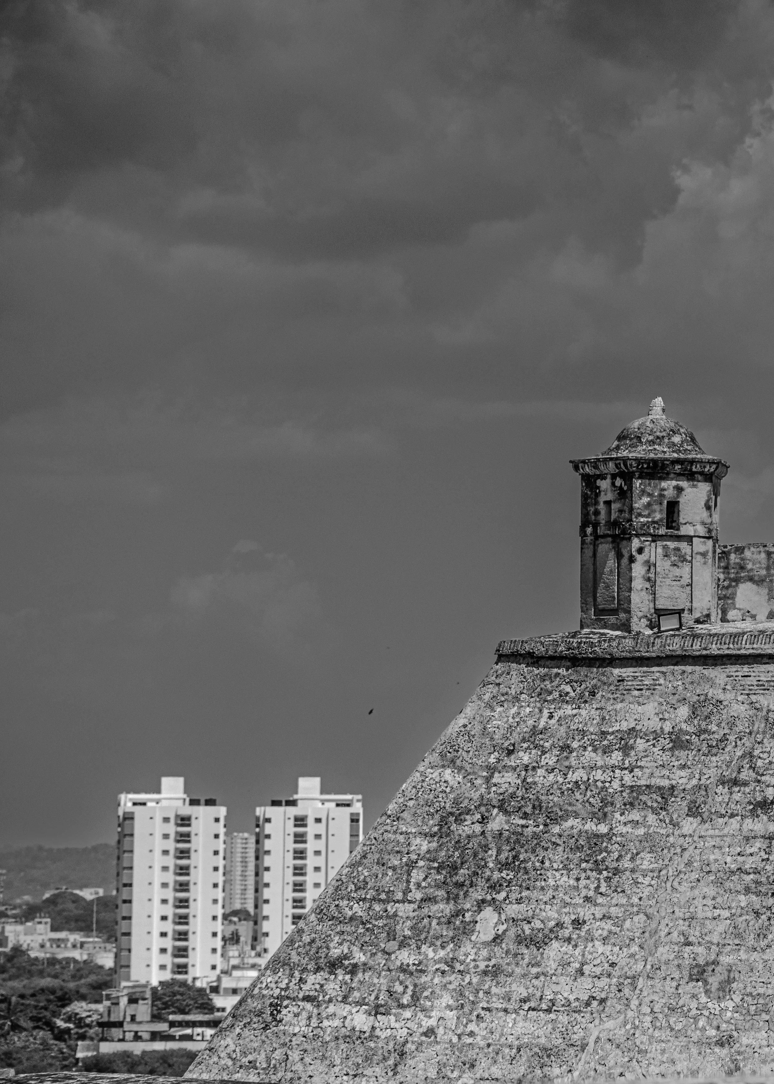 Black and white photo of a historic stone tower on a sloped roof, with modern high-rise buildings in the background under cloudy sky.