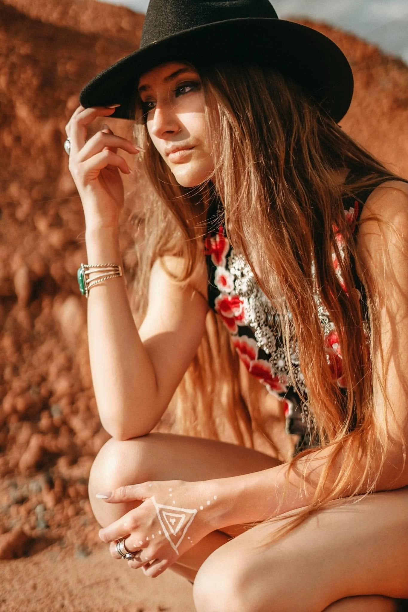 A woman in a black hat and floral dress, sitting on the ground with a natural background, gazing thoughtfully.