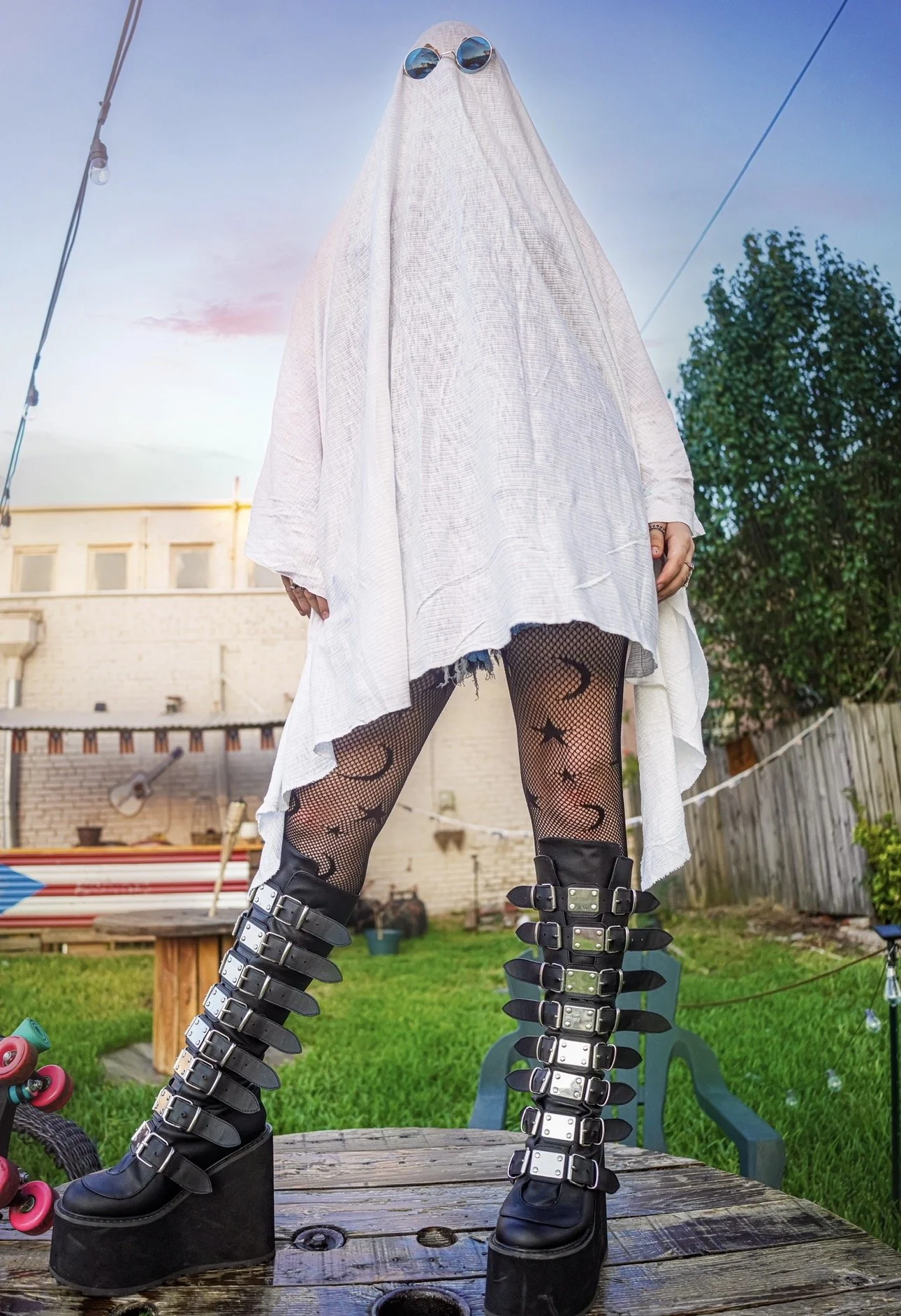 Person standing outdoors on a table wearing black platform boots with multiple buckle straps, fishnet tights with moon and star designs, a white oversized shirt, and sunglasses, with a garden and building in the background.