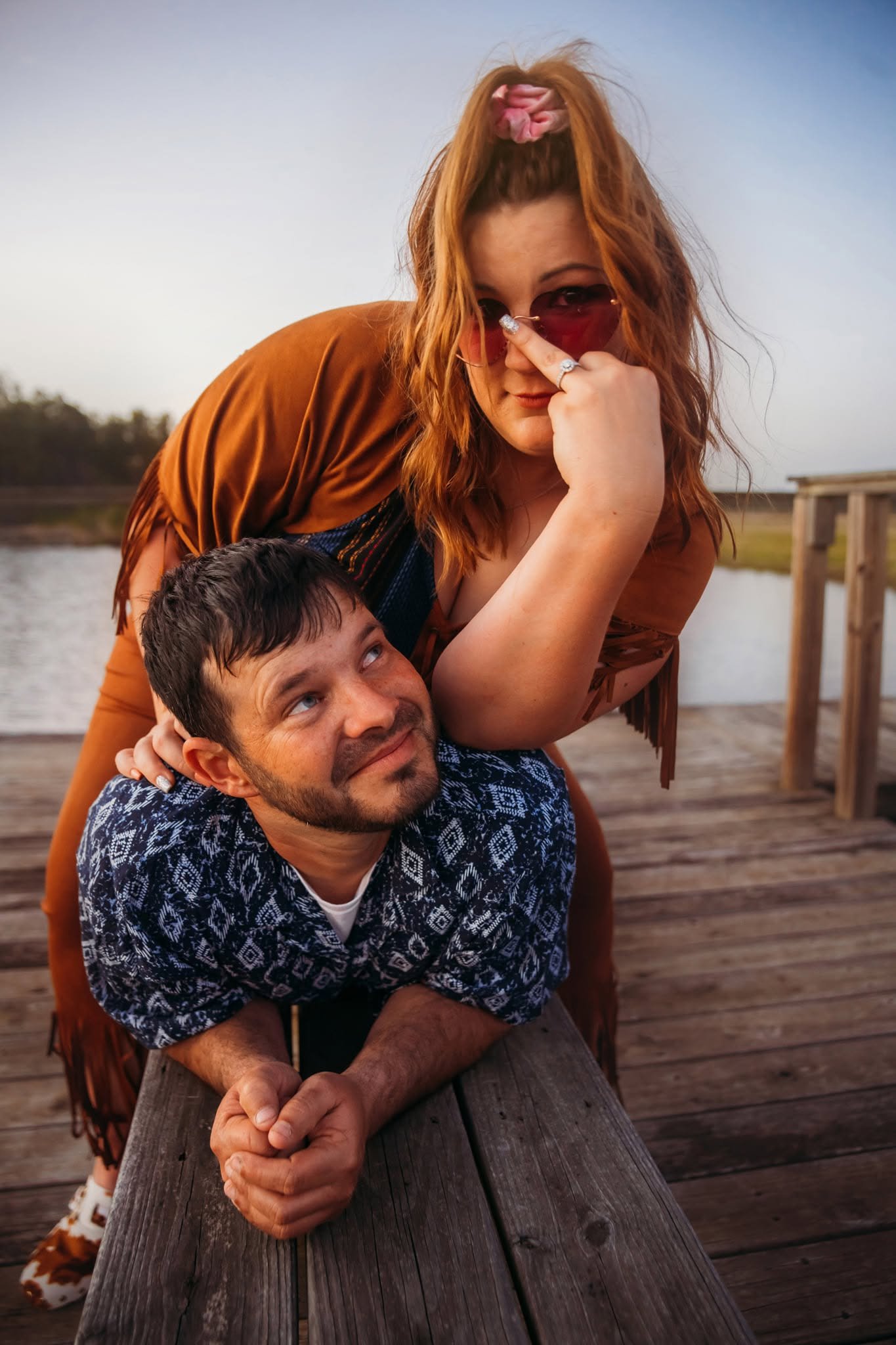 A woman with long red wavy hair, wearing red-tinted sunglasses, a brown jacket, and a scrunchie on her head, is leaning above a man with short dark hair and a beard. The man is sitting at a wooden table outdoors by a body of water, with trees and a cloudy sky in the background. The woman is playfully pointing at her sunglasses while the man looks up at her with a smile.