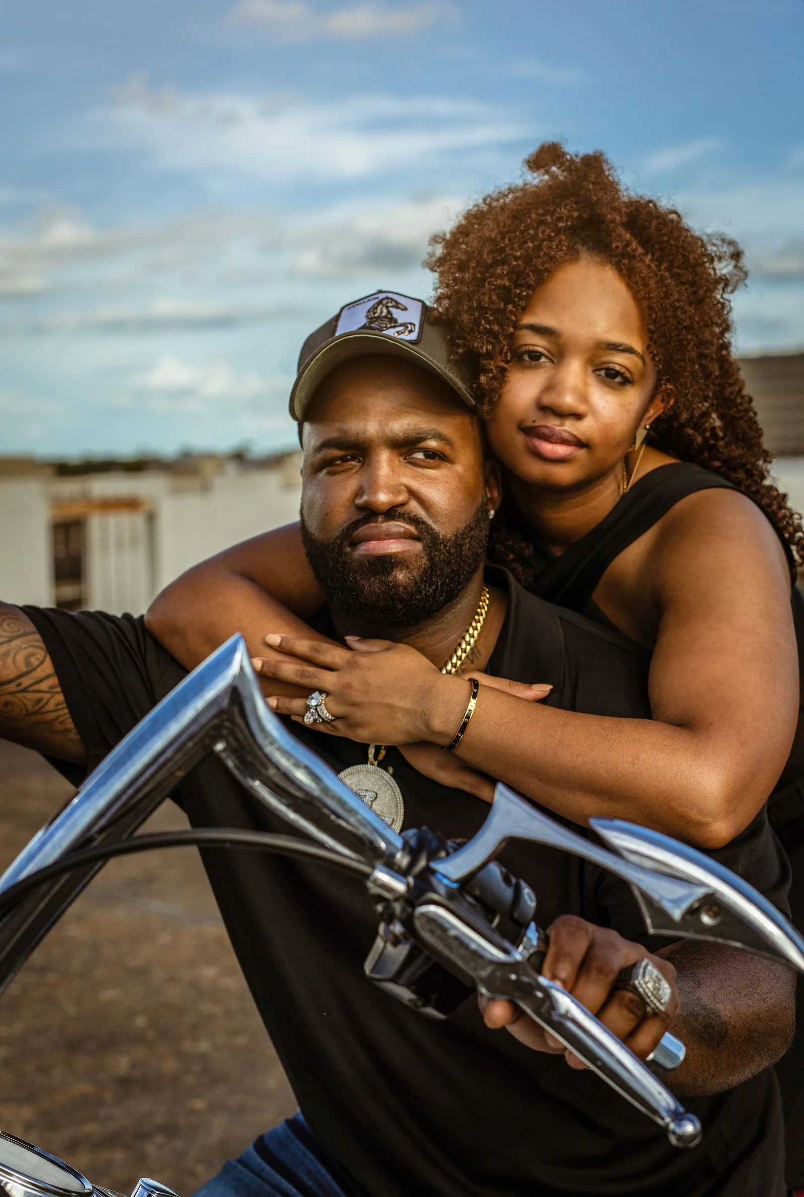 A man and woman sitting on a motorcycle together outdoors during daytime, with a background of buildings and a partly cloudy sky.