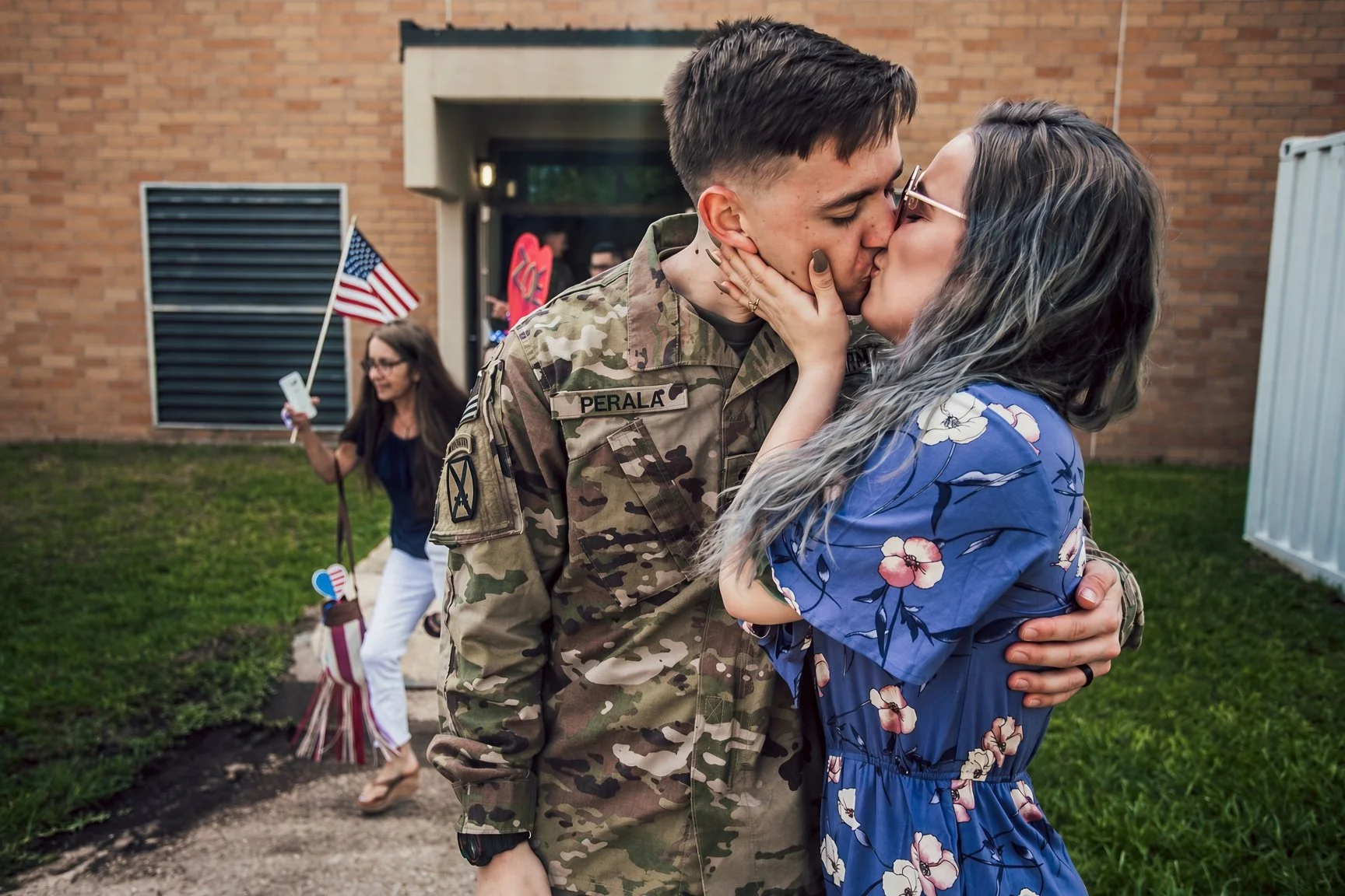 A soldier in uniform kissing a woman in a blue floral dress outside a building, with a woman holding an American flag in the background.