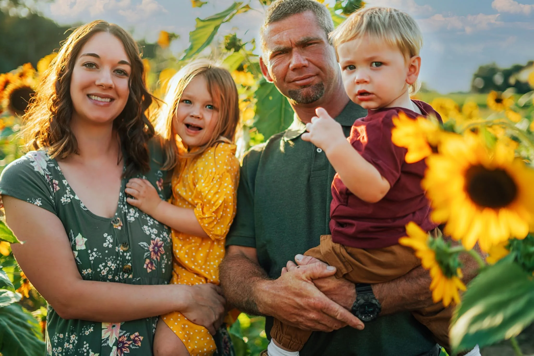 A family of five posing outdoors among sunflowers in a sunflower field during daytime. The family includes a woman with curly brown hair, two young girls, a man with short gray hair and beard, and a young boy with blonde hair. The woman is smiling, the girls are looking at the camera with one smiling and the other with a surprised expression, the man looks slightly puzzled or serious, and the boy is looking at something off-camera with a concerned expression.
