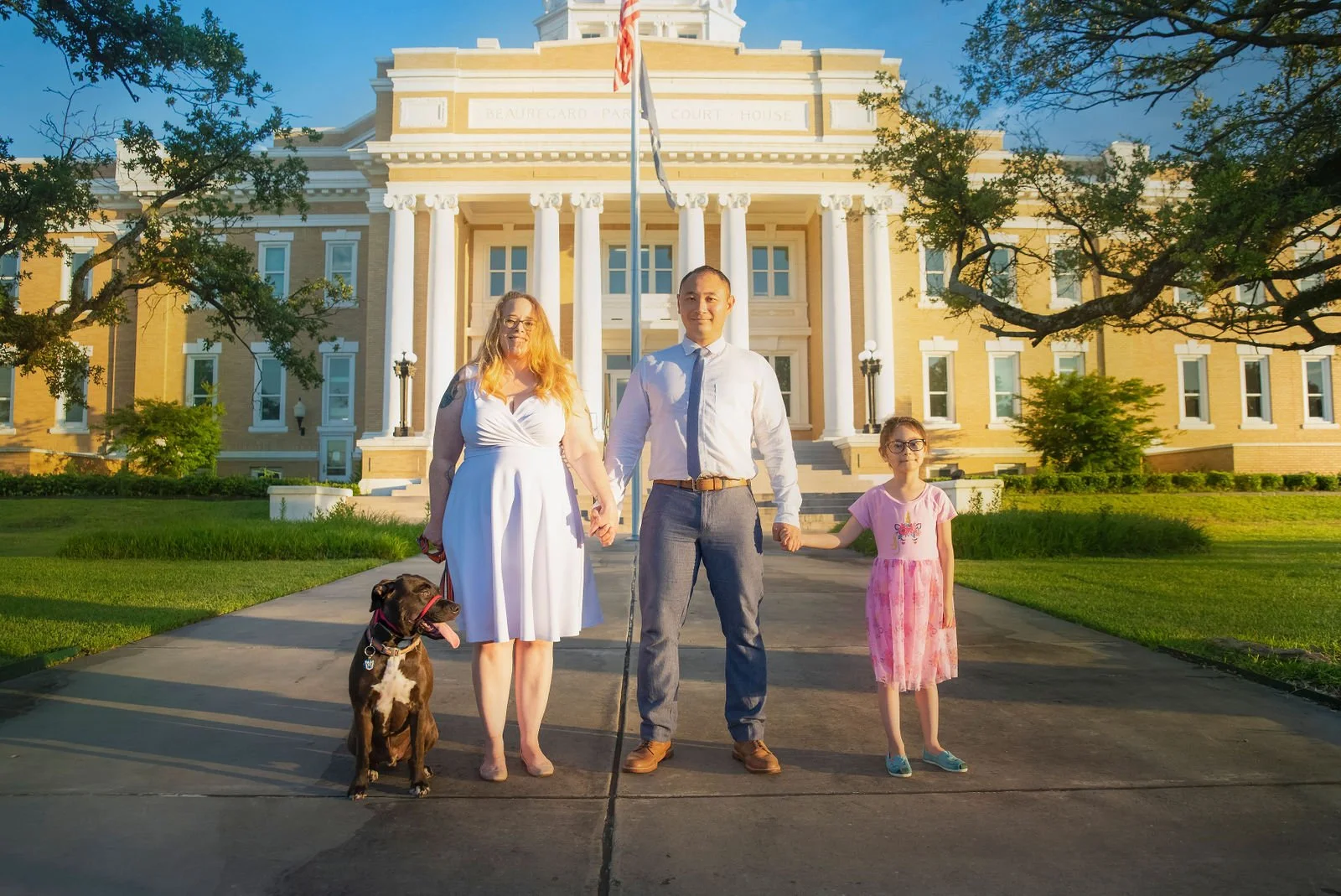 A family of three, a woman, a man, and a young girl, standing in front of a large, historic courthouse with columns, holding hands, with a dog sitting beside the woman, during late afternoon or early evening, with trees and a well-maintained lawn surrounding them.