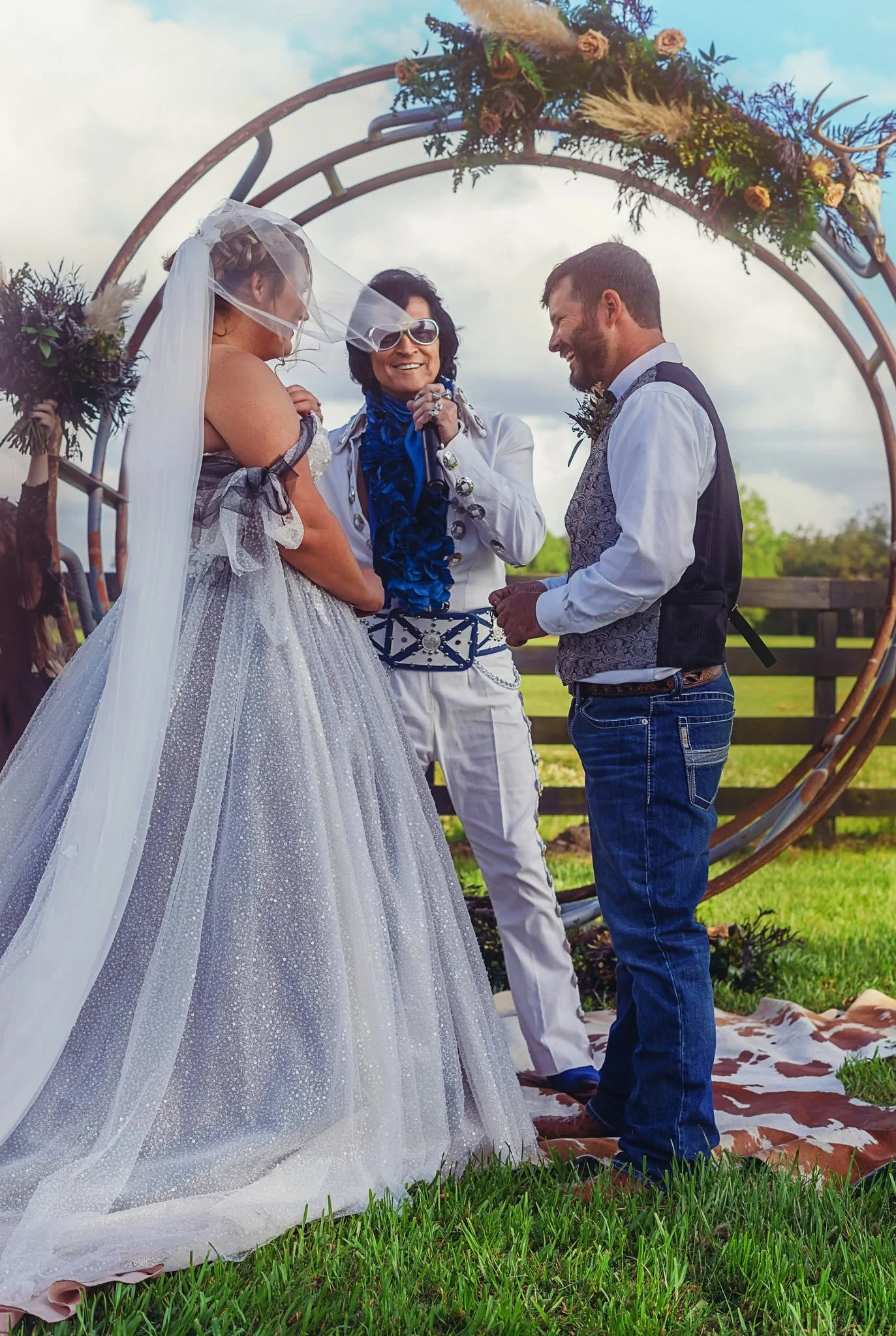 A wedding ceremony outdoors with a bride in a sparkly white wedding dress and veil, a groom in a vest and jeans, and a woman officiant in white with sunglasses, all standing under a circular floral arch with pink roses and greenery. They are smiling and holding hands, on a grassy field with a wooden fence and trees in the background.