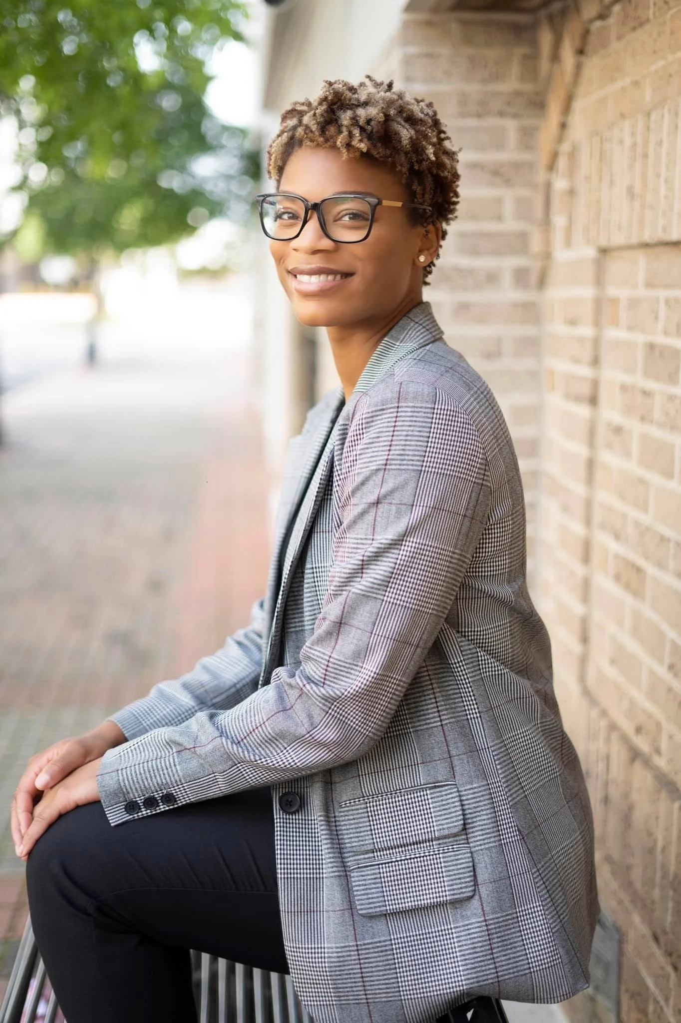 A woman with short curly hair wearing glasses, a gray plaid blazer, and black pants sitting outdoors near a brick wall, smiling at the camera.