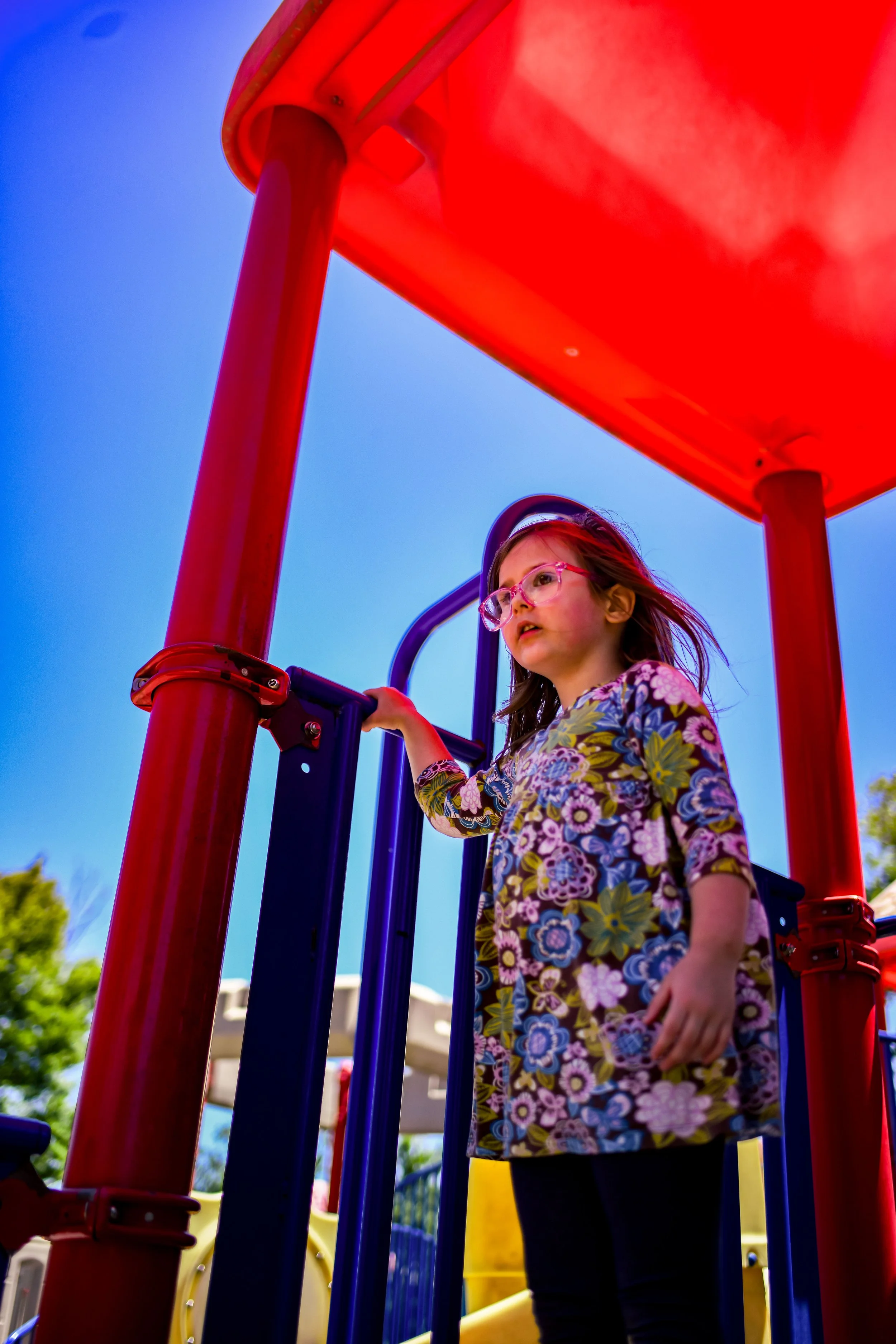 A young girl wearing glasses and a floral shirt, standing on a playground beneath a red shade structure, with blue sky and trees in the background.