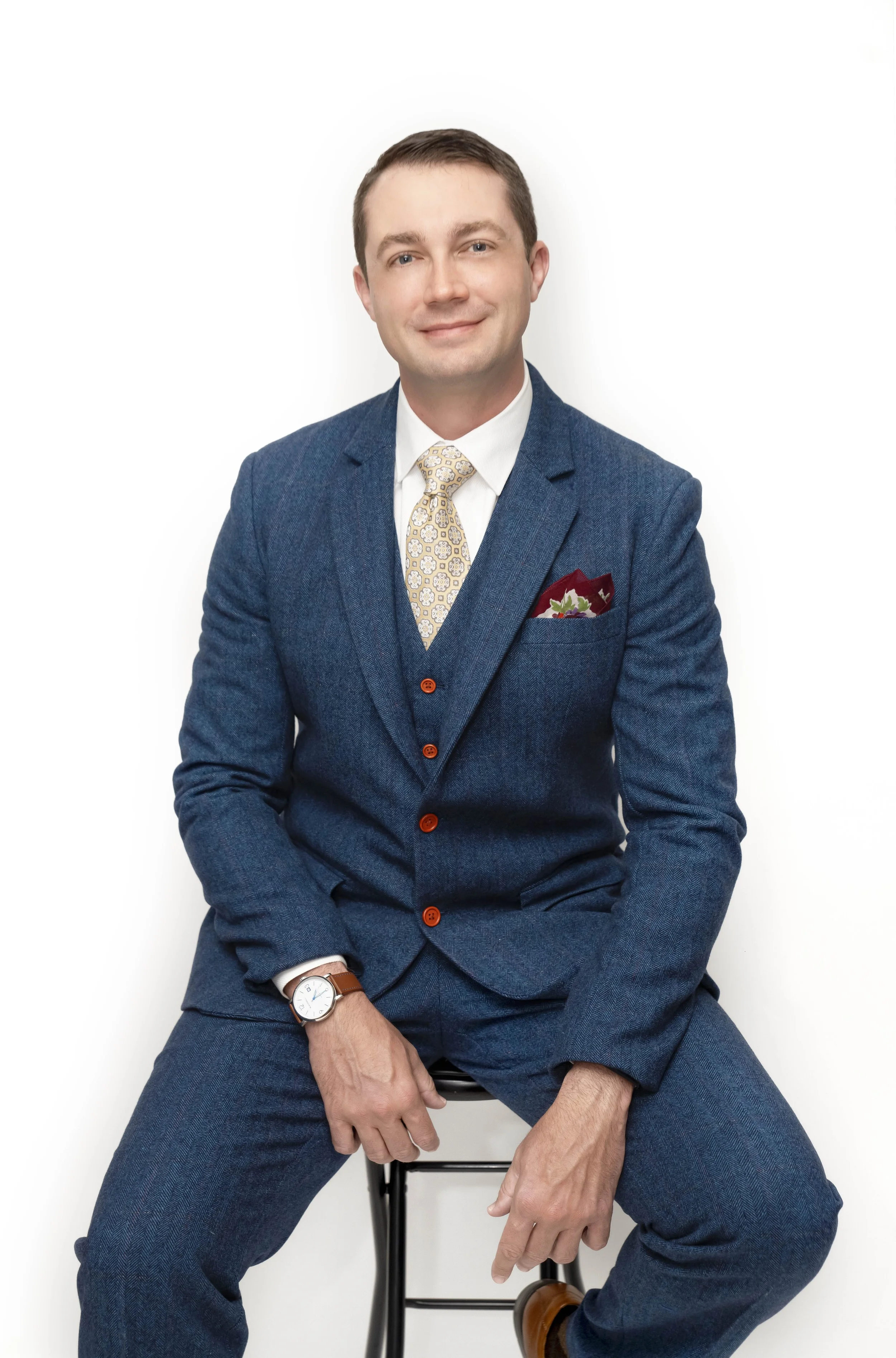A man wearing a dark blue suit, yellow patterned tie, white shirt, and brown shoes sitting on a chair, with arms crossed, positioned against a plain white background.