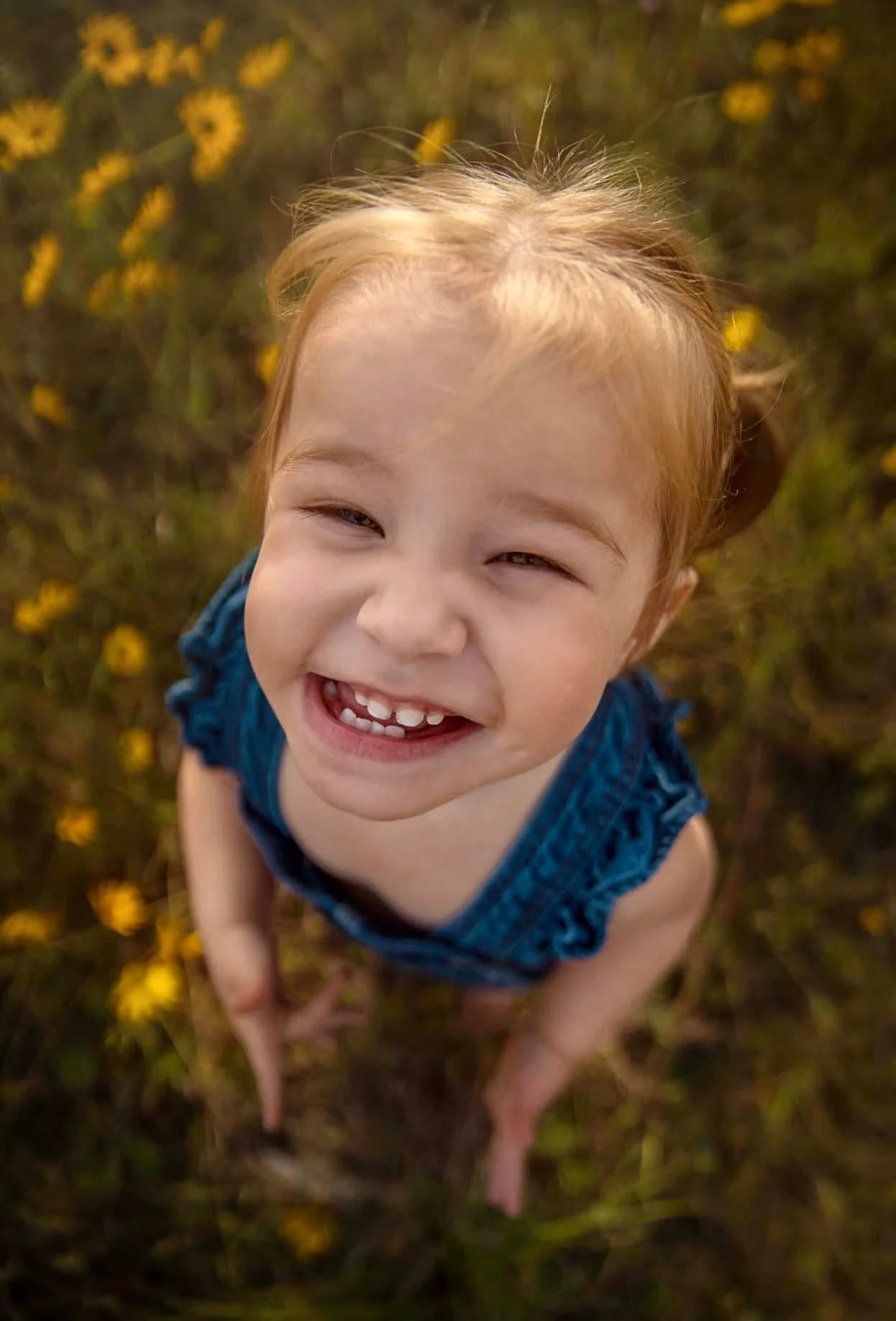 A young girl with red hair, smiling and looking up at the camera, standing amidst yellow flowers.