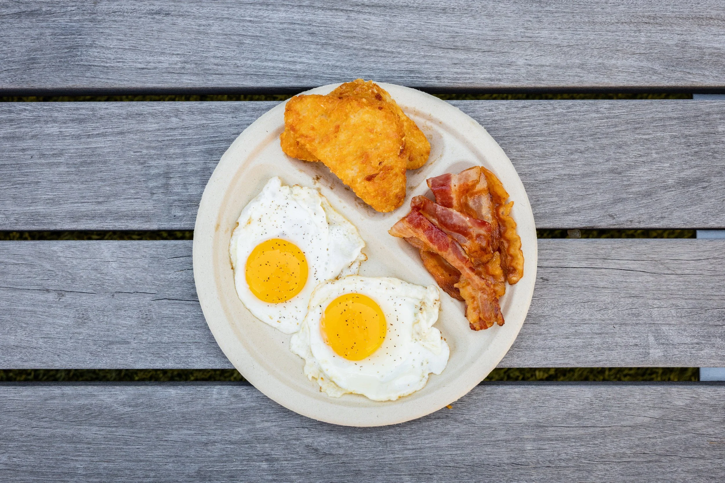 Breakfast plate with two sunny side up eggs, bacon strips, a piece of fried chicken, and a piece of ham, placed on a wooden table.