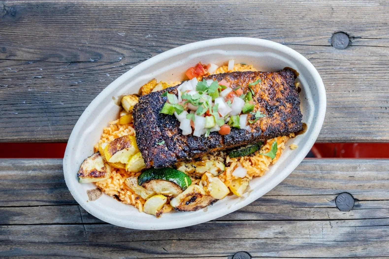 Grilled fish fillet served on bed of seasoned rice with roasted vegetables and topped with chopped onions, tomatoes, and cilantro, in a disposable oval plate on a wooden surface.