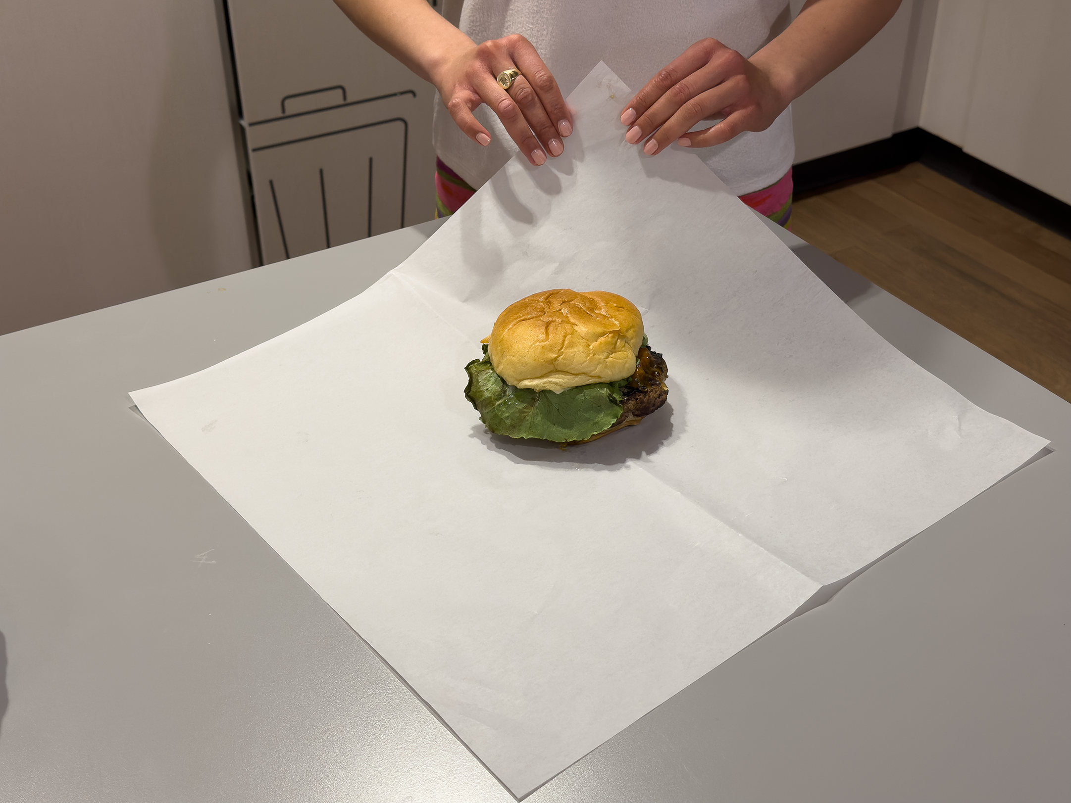 Person tearing paper back from a sandwich. The sandwich has a bun, lettuce, and cooked meat, placed on white parchment paper on a gray table.