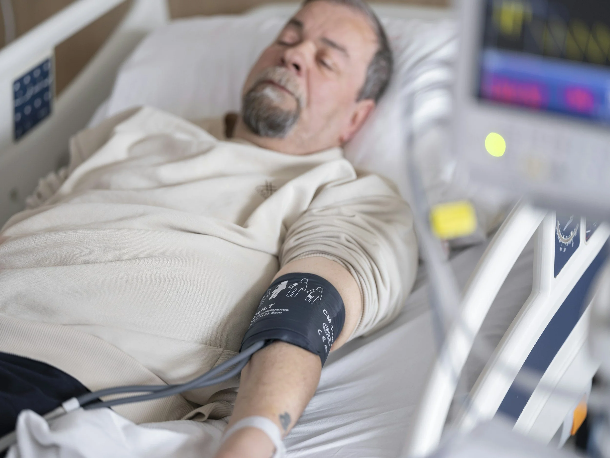 A man lying in a hospital bed with his eyes closed, connected to medical monitoring devices, wearing a dark blood pressure cuff on his arm.