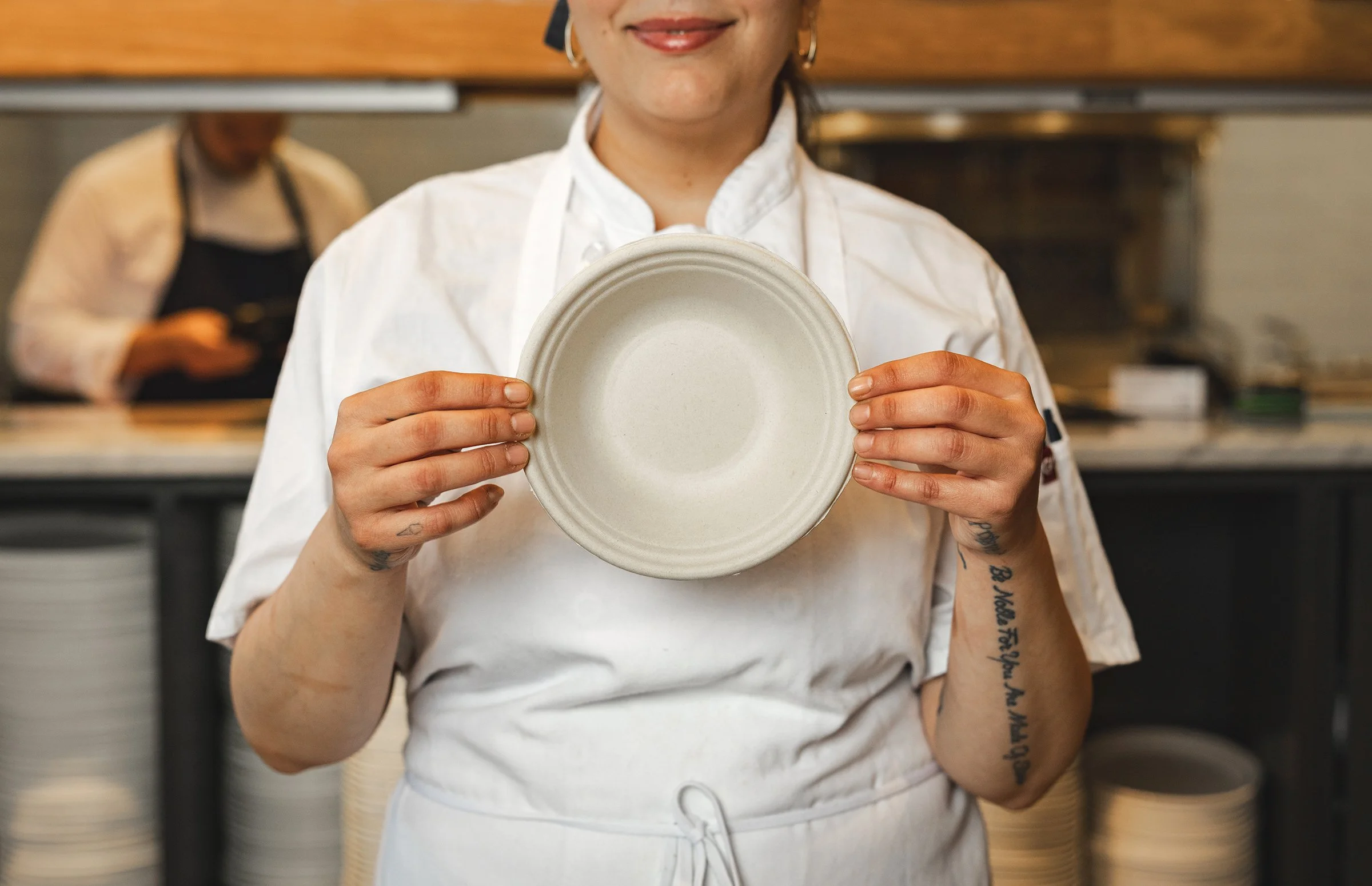 A woman in a white chef's coat holding an empty beige ceramic plate in a commercial kitchen, with another person in the background wearing a black apron